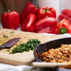 Photo of a meal being prepared with chopped herbs on a cutting board, ground meat in a frying pan, and whole red peppers in the background